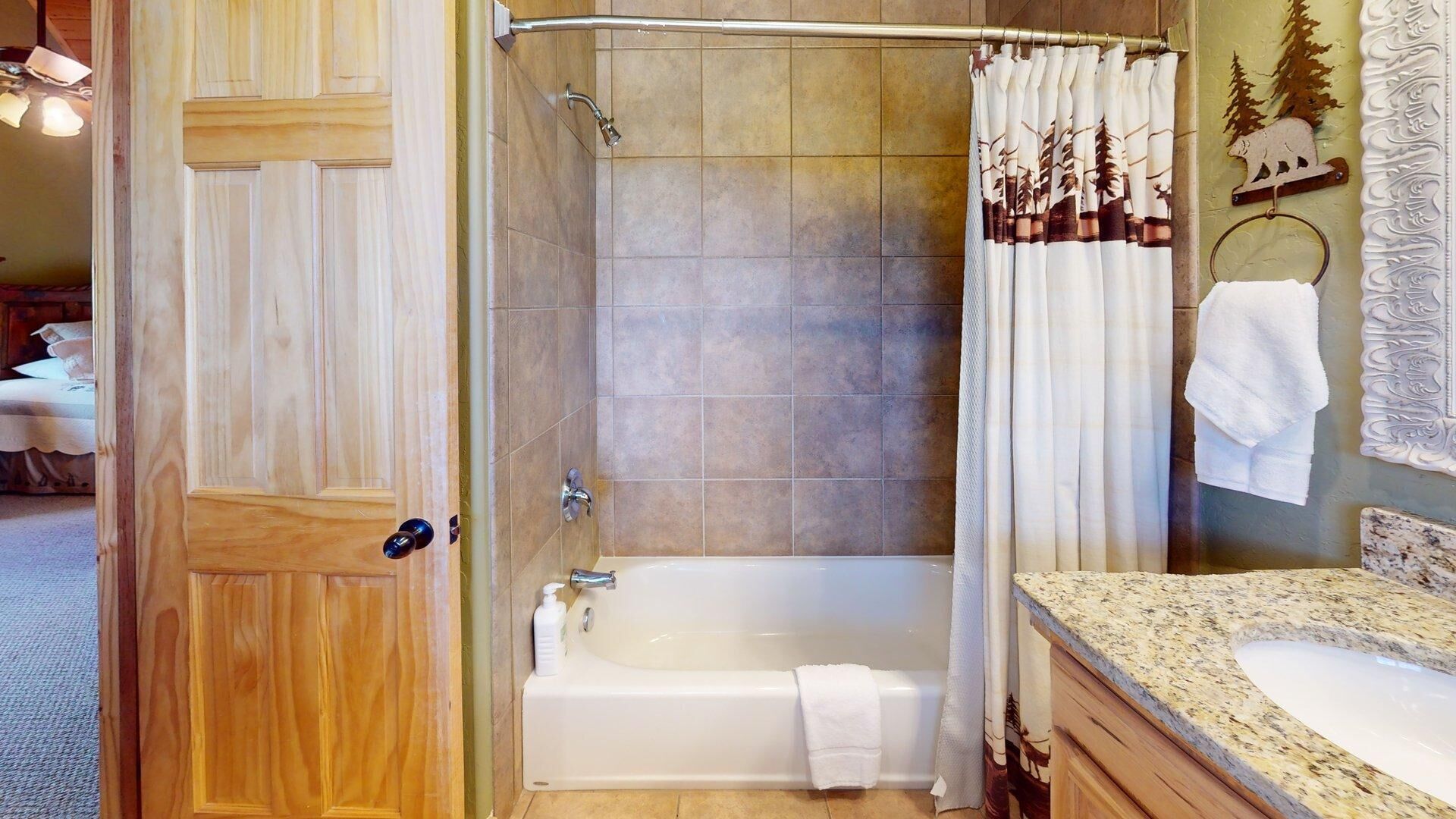 A bathroom with a wooden door, a bathtub, a shower curtain with a forest design, and a granite countertop.