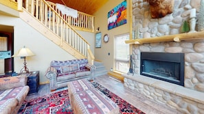 A cozy living room with a stone fireplace, a staircase, and a colorful abstract painting. - Master ensuite | Wood burning fireplace | Bike & Ski Gear Storage | Byrd's Nest (Angel Fire)
