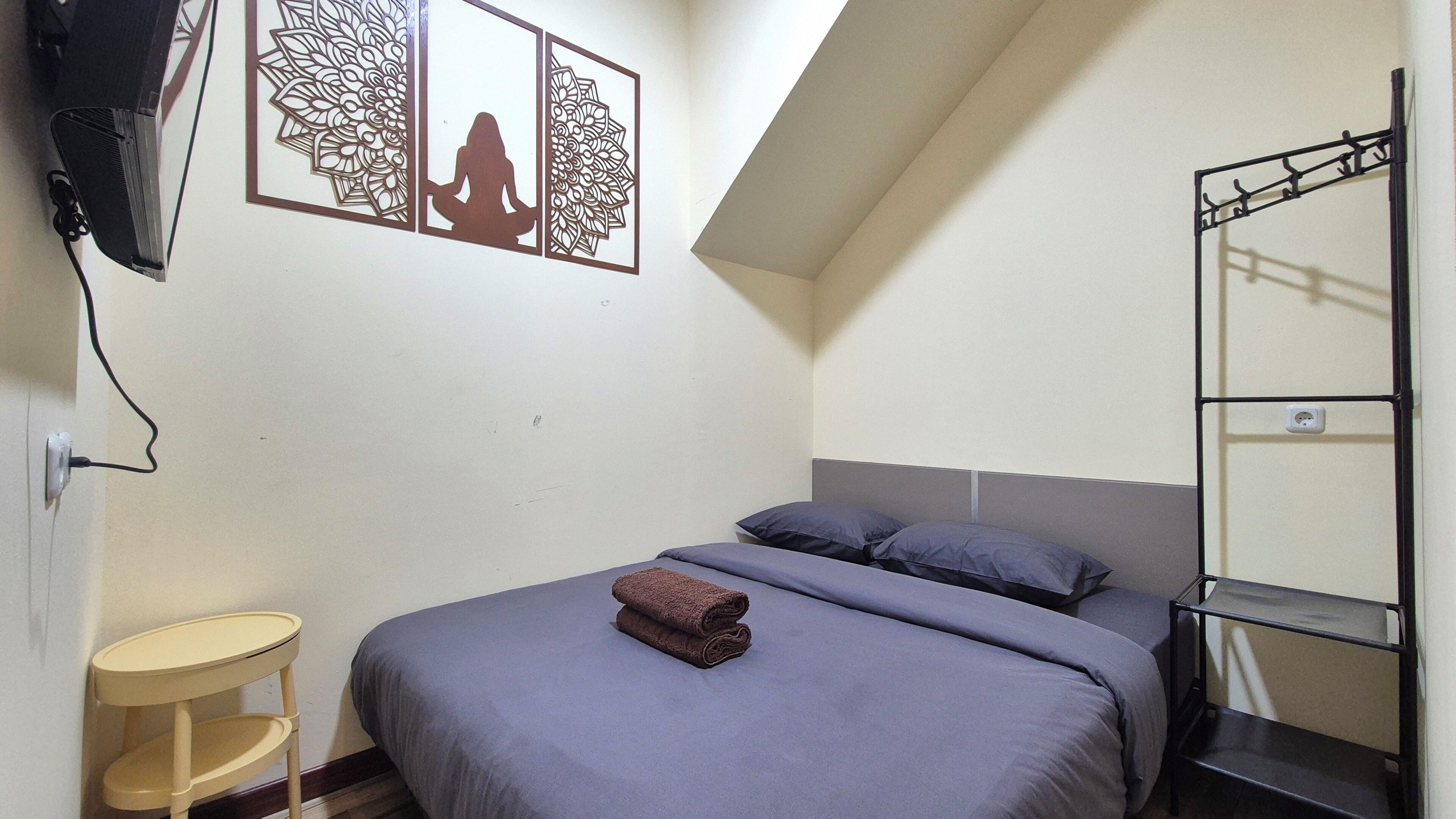 Un dormitorio con cama, almohadas, una mesita, un televisor montado en la pared y un panel decorativo.