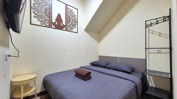 Un dormitorio con cama, almohadas, una mesita, un televisor montado en la pared y un panel decorativo.