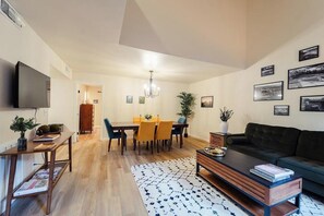 A modern living room with a flat-screen TV, a dark sofa, a coffee table, and a dining area with a table and chairs. - Goldies Birdie Roost (Hot Springs Village)