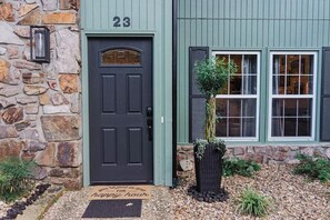 Une maison avec une façade en pierre, une porte vert foncé portant le numéro 23 et un paillasson avec un message.
