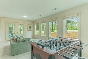 A room with a foosball table, a sofa, and large windows with a view of trees. - Pepper's Pines and Putts, on Isabella golf course (Hot Springs Village)