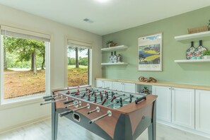 Um quarto com mesa de pebolim, janela com vista para árvores e parede com quadro e prateleiras.