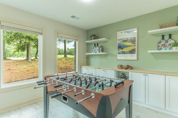 A room with a foosball table, a window with a view of trees, and a wall with a framed picture and shelves. - Pepper's Pines and Putts, on Isabella golf course (Hot Springs Village)
