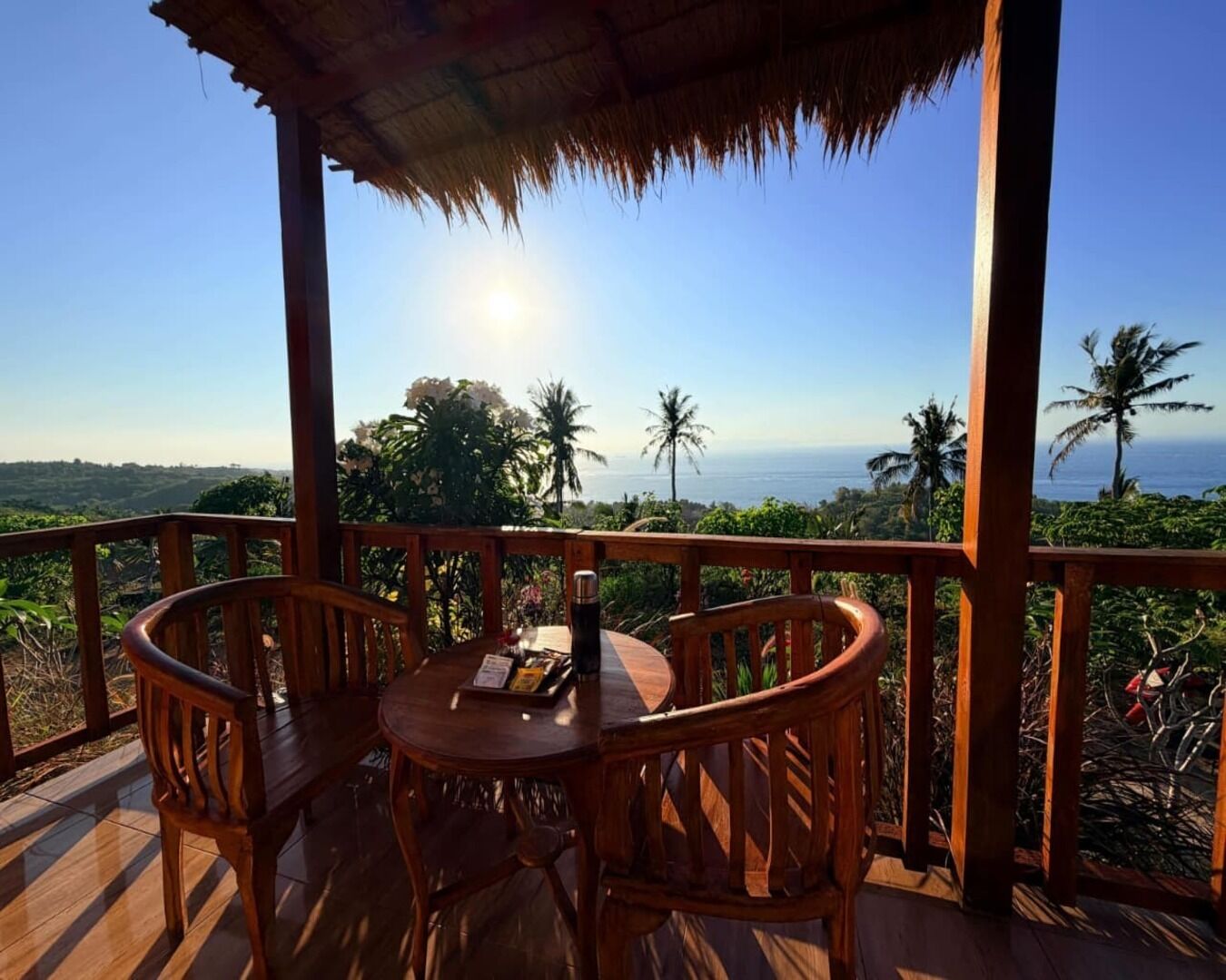 Eine Holzterrasse mit Tisch und Stühlen, mit Blick auf eine Küstenlandschaft mit Palmen und dem Meer.