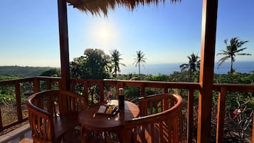 Eine Holzterrasse mit Tisch und Stühlen, mit Blick auf eine Küstenlandschaft mit Palmen und dem Meer.