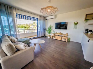 A modern living room with a sofa, a small round table, a TV mounted on the wall, and a balcony with a view. - Teani Skyline | Downtown Papeete • Terrace • Moorea View • A/C • Parking (Papeete)