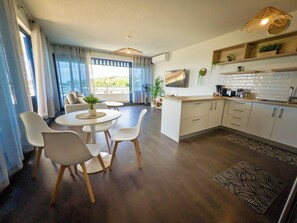A modern kitchen with white cabinets, a dining area with a round table and chairs, and a living area with a sofa and a TV. - Teani Skyline | Downtown Papeete • Terrace • Moorea View • A/C • Parking (Papeete)