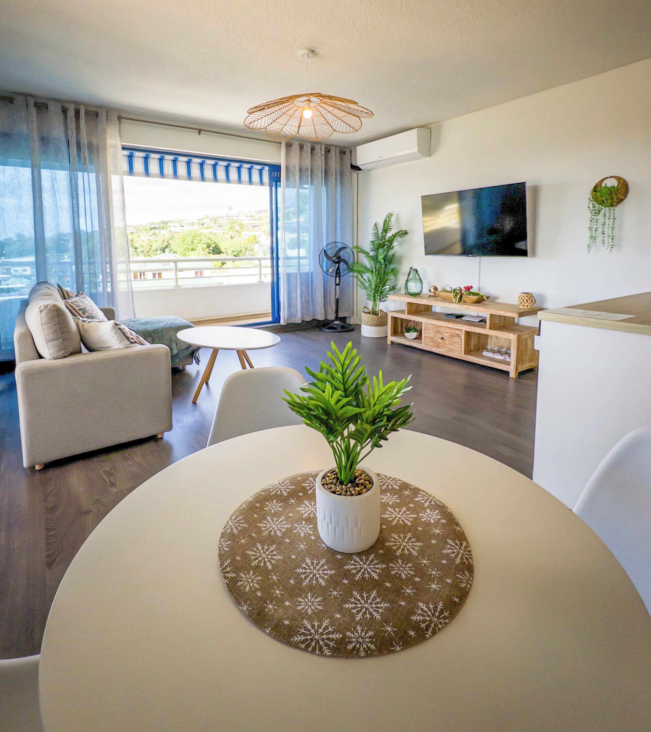 Un salon moderne avec un canapé, une table à manger agrémentée d’un plateau décoratif, une plante en pot et un téléviseur fixé au mur.
