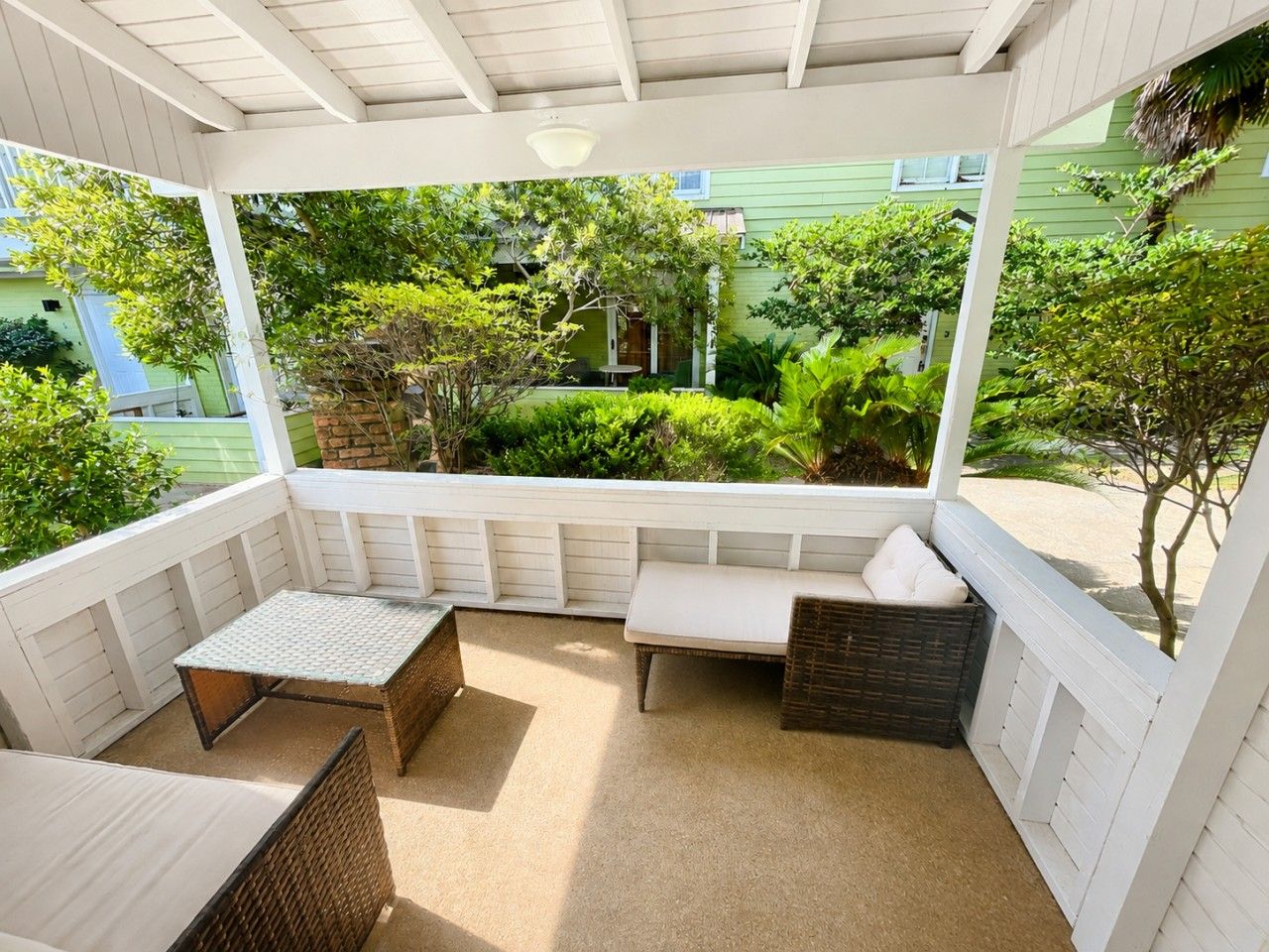 A covered outdoor seating area with a bench and a table, surrounded by greenery and a building in the background.