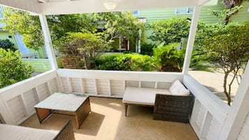 A covered outdoor seating area with a bench and a table, surrounded by greenery and a building in the background.