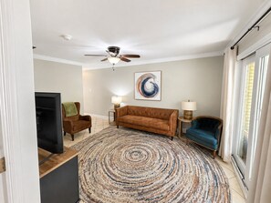 A living room with a brown sofa, a blue armchair, a patterned rug, and a ceiling fan. - Palm & Pearl | Walk to Beach & Downtown (Ocean Springs)