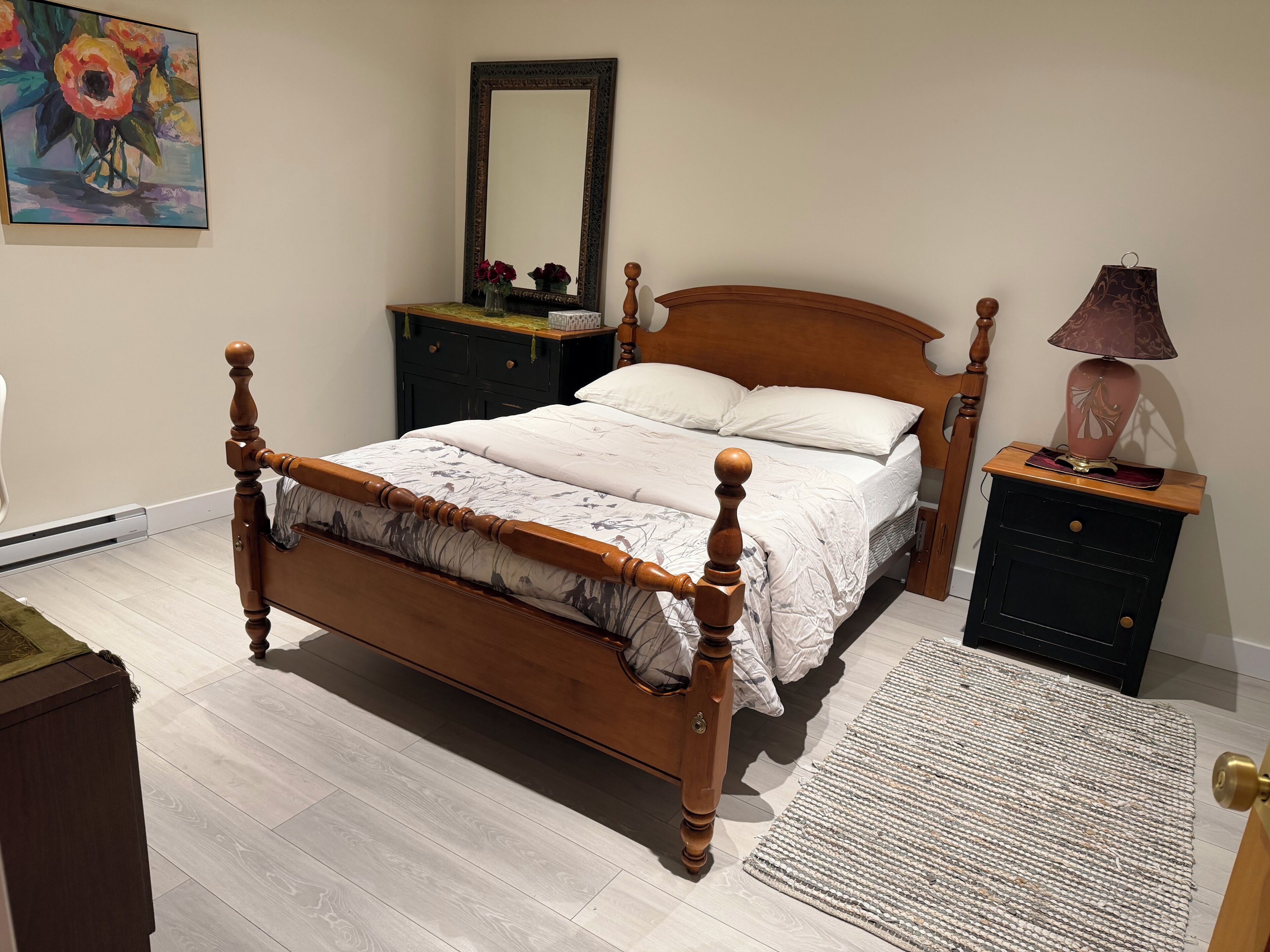 A bedroom with a wooden four-poster bed, a nightstand with a lamp, a dresser, a mirror, and a painting on the wall.