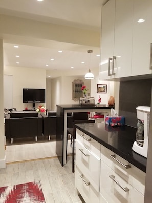 A modern kitchen with white cabinets, a black countertop, and a dining area with a black sofa and a TV. - North Van Escape: Private 2BR Basement Near Grouse & Capilano Bridge (North Vancouver)