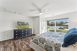 A bedroom with a bed, dresser, ceiling fan, and a window with a view of a lake and buildings.