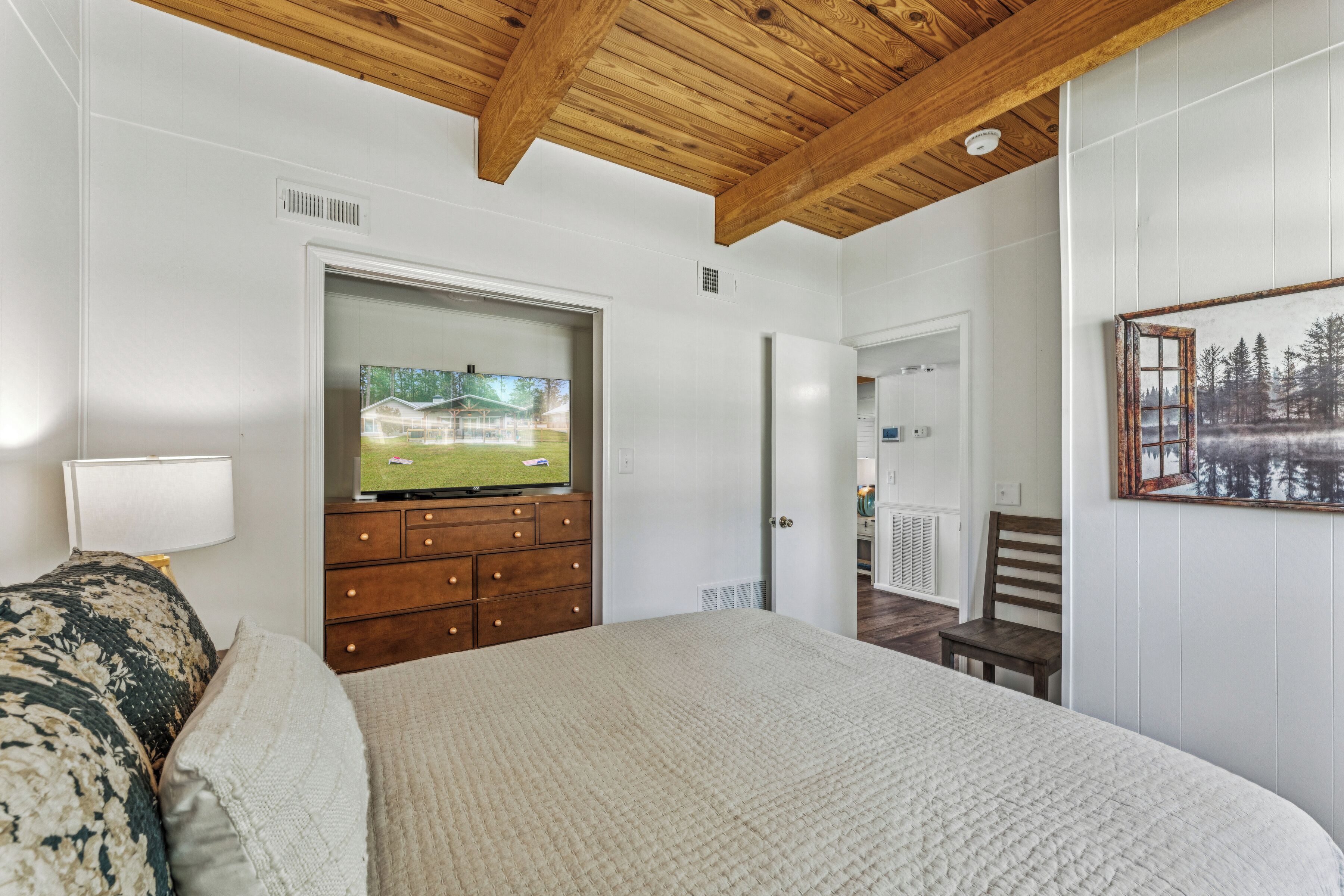 Une chambre à coucher avec un grand lit, des poutres en bois au plafond, un téléviseur posé sur une commode et une porte donnant sur une autre pièce.