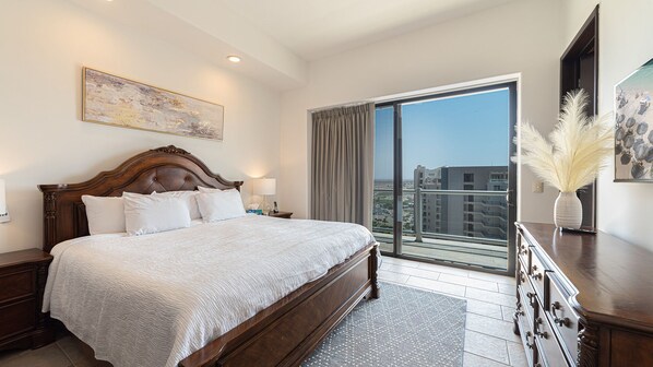 Ein Hotelzimmer mit einem großen Bett, einem Nachttisch, einer Kommode, einem Bild an der Wand und Blick auf die Stadt durch ein großes Fenster.