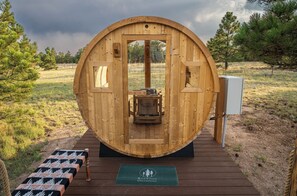 A wooden structure with a circular door, a small table with a chair, and a sign with a logo. - Wild Finch Stay - Sauna • Cold Plunge • Hot Tub (Colorado Springs)