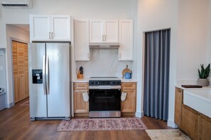 A modern kitchen with white cabinets, a stainless steel refrigerator, a built-in oven, and a grey curtain. - Wild Finch Stay - Sauna • Cold Plunge • Hot Tub (Colorado Springs)