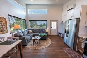 A modern kitchen with a built-in refrigerator, a dining area with a table and chairs, a gray sofa, and a window with blinds. - Wild Finch Stay - Sauna • Cold Plunge • Hot Tub (Colorado Springs)