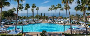 A large pool surrounded by lounge chairs and umbrellas, with palm trees and a view of the ocean.