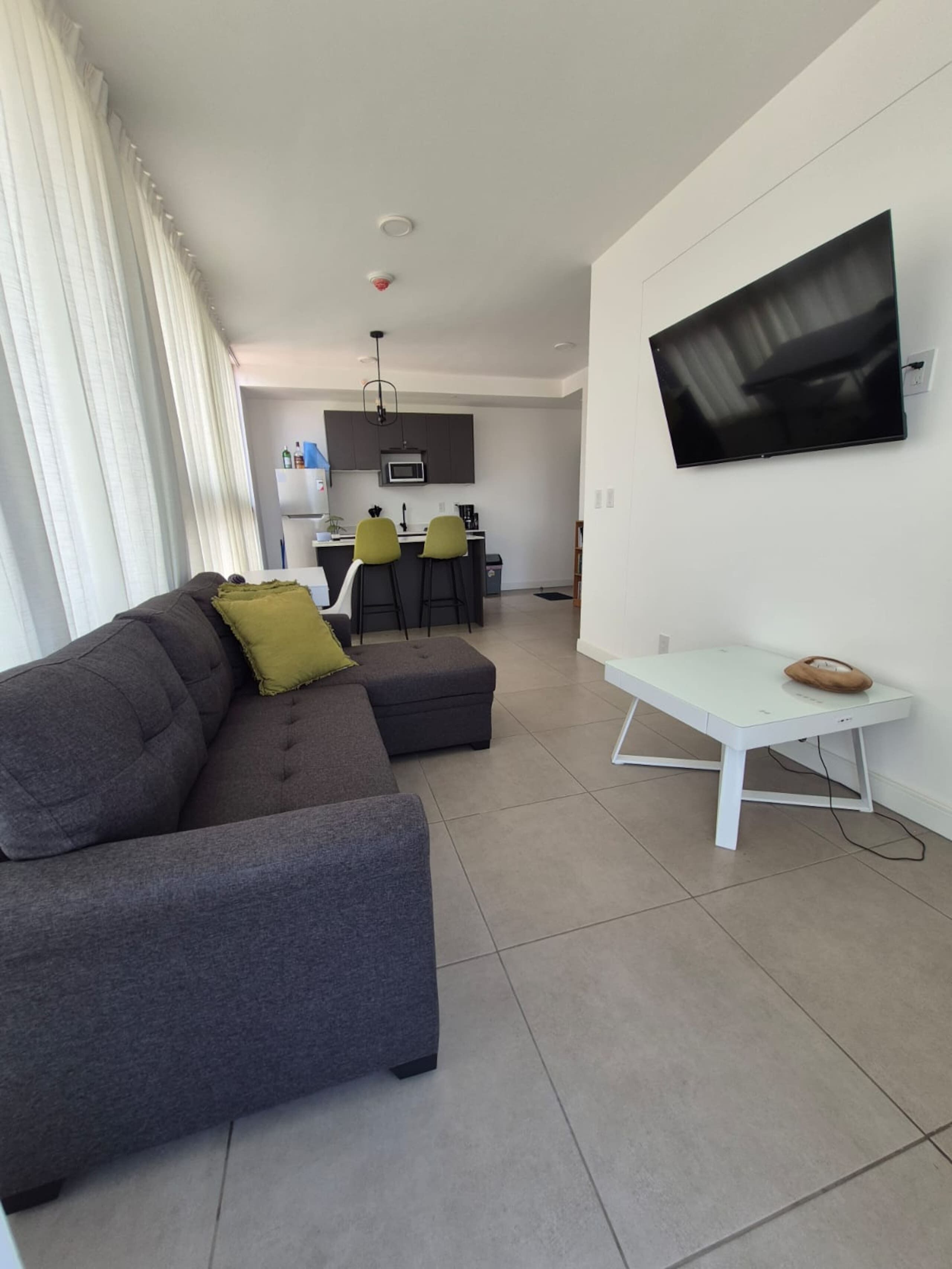 A modern living room with a grey sofa, a small white table, and a flat-screen TV mounted on the wall.