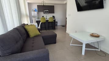 A modern living room with a grey sofa, a small white table, and a flat-screen TV mounted on the wall.