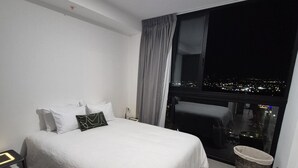 A hotel room with a bed, a nightstand, a window with a city view, a window with curtains, and a small trash bin. - Liva Escalante Sky Retreat | Panoramic City Views & Walking Distance to Restaurants (San José)