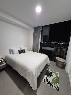 Ein Hotelzimmer mit einem Bett, einem Nachttisch, einem Fenster mit Stadtblick, einem Vorhangfenster und einer kleinen Mülltonne.