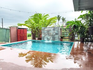 Una piscina rectangular con una cascada, rodeada de abundante vegetación y una cerca roja.