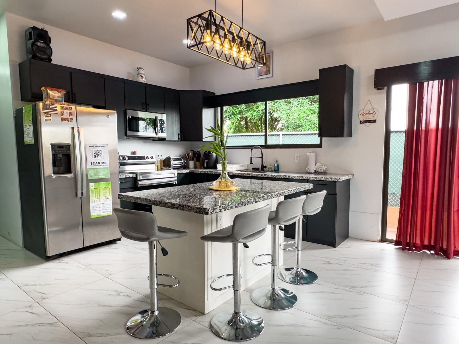 A modern kitchen with a central island, bar stools, a refrigerator, microwave, and a large window with a view of greenery.