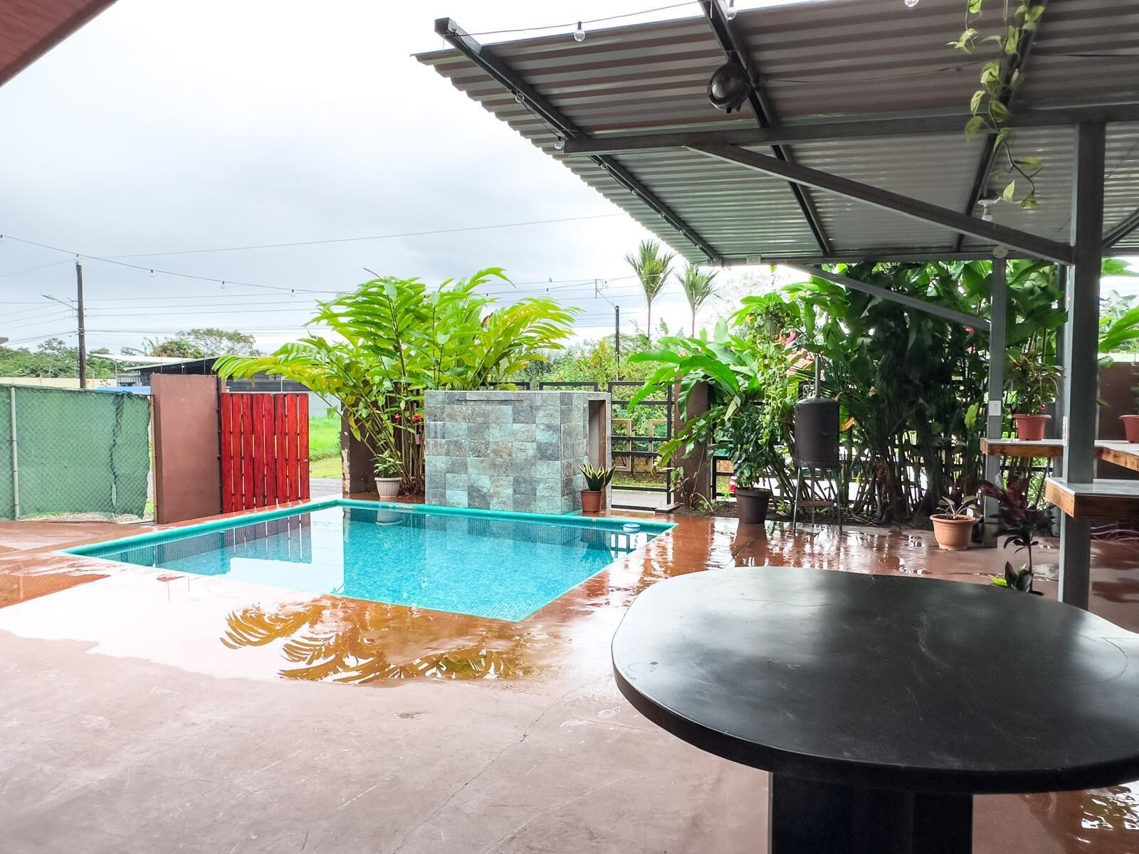 A swimming pool with a tiled deck, surrounded by potted plants and a covered patio area.