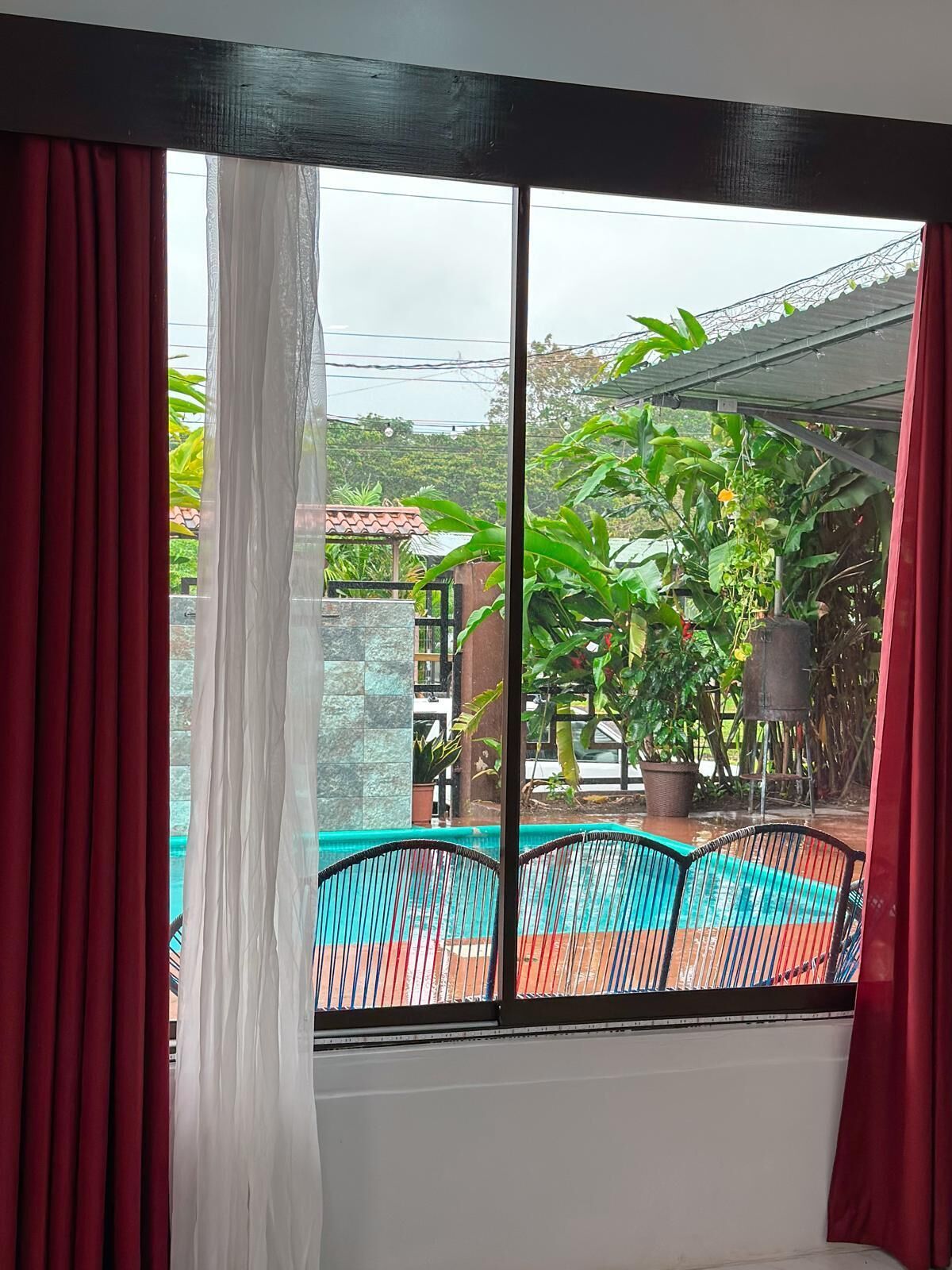 A room with a large window, a view of a swimming pool, and a covered outdoor area with plants.