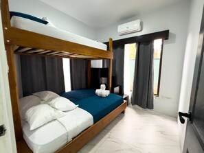 Habitación con literas, colchón azul, almohadas blancas y cabecero de madera. Cuenta con una ventana con cortinas, un equipo de aire acondicionado y una puerta.