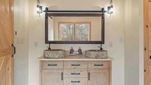 A bathroom with a wooden vanity, two round mirrors, a large mirror above the vanity, and a window with a view of trees. - Winter Court Log Cabin by Avanstay Private Trail + Fire Pit + Basketball Court Near Camelback (Pocono Summit)