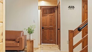 A hallway with a wooden door, a brown leather sofa, a small wooden table with a plant, and a wooden staircase. - Winter Court Log Cabin by Avanstay Private Trail + Fire Pit + Basketball Court Near Camelback (Pocono Summit)