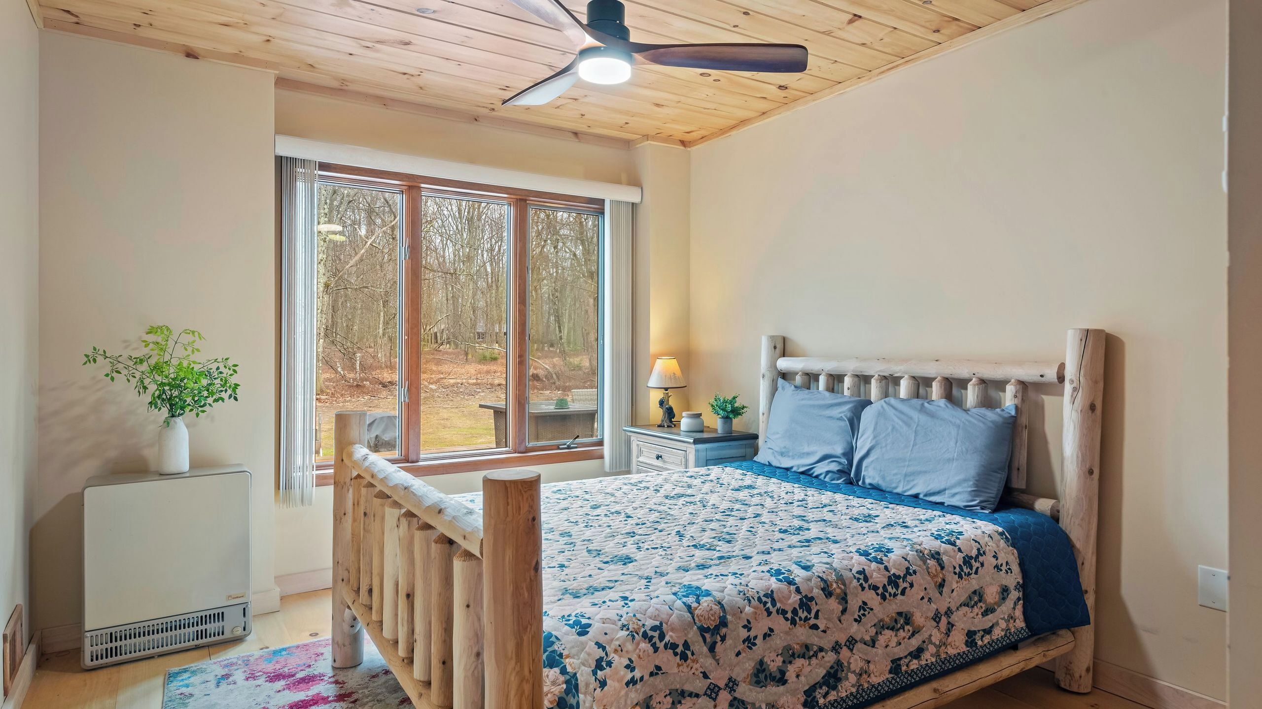 A bedroom with a wooden bed, a nightstand, a lamp, a window with a view of trees, and a ceiling fan.