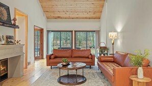 A spacious living room with a fireplace, leather sofas, a round coffee table, and large windows offering a view of the outdoors. - Winter Court Log Cabin by Avanstay Private Trail + Fire Pit + Basketball Court Near Camelback (Pocono Summit)