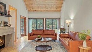 A spacious living room with a fireplace, leather sofas, a round coffee table, and large windows offering a view of the outdoors. - Winter Court Log Cabin by Avanstay Private Trail + Fire Pit + Basketball Court Near Camelback (Pocono Summit)