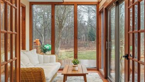 A room with a white sofa, a wooden table, and a view of a forest through large glass doors. - Winter Court Log Cabin by Avanstay Private Trail + Fire Pit + Basketball Court Near Camelback (Pocono Summit)