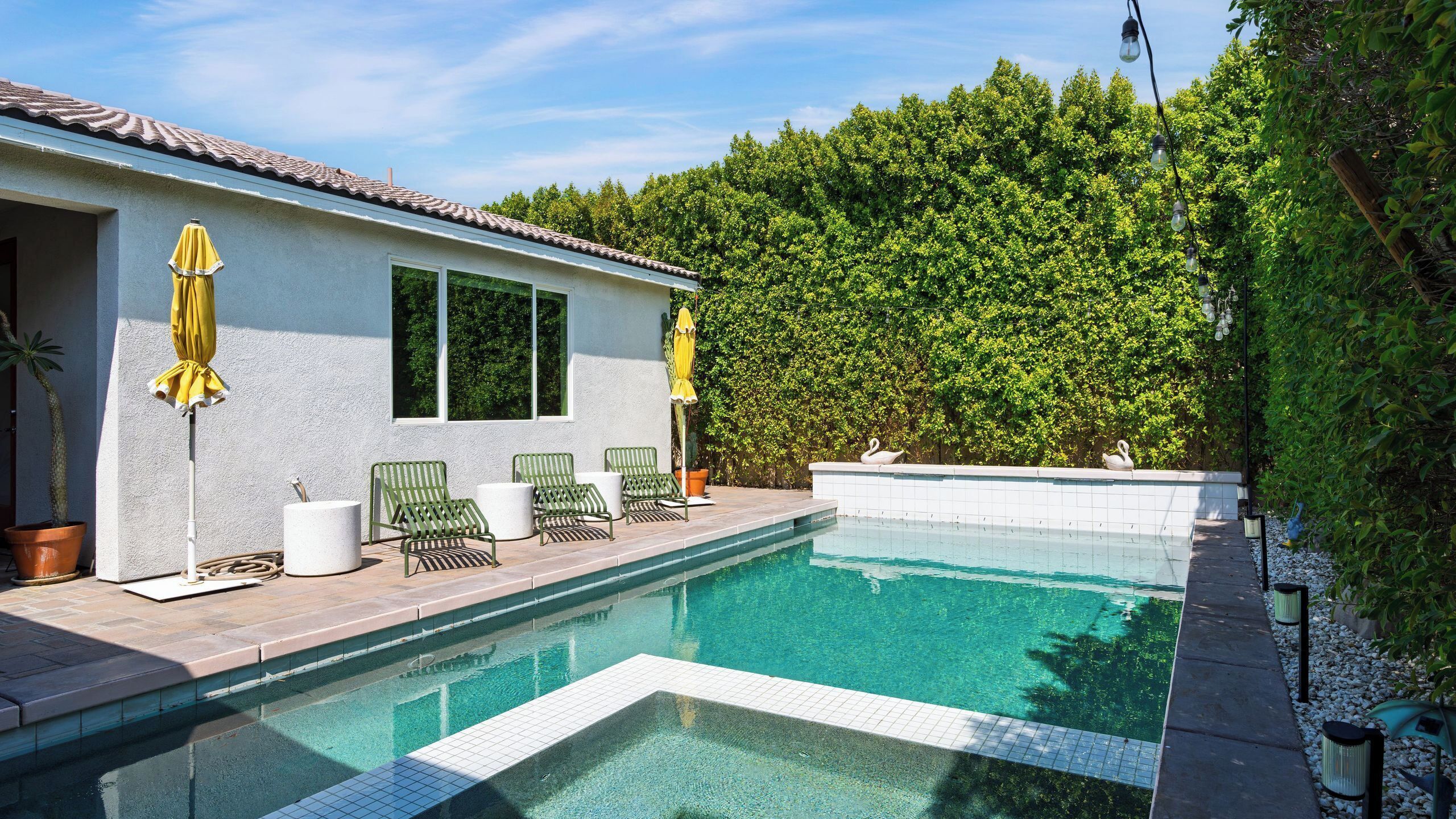 A swimming pool with a tiled deck, two green lounge chairs, and a white wall with a window.
