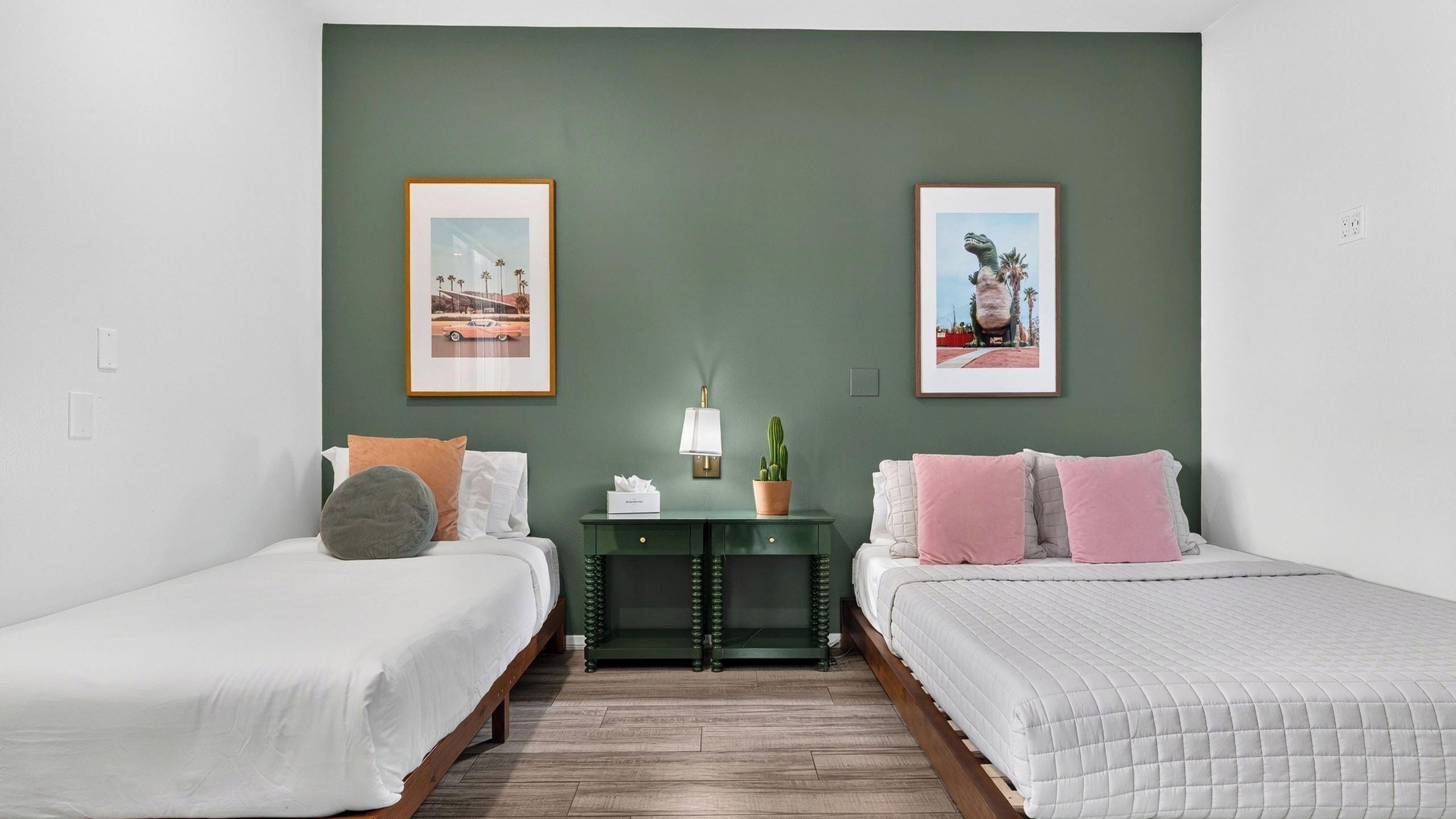 Ein Hotelzimmer mit zwei Einzelbetten, einem Nachttisch mit Lampe und Blumentopf sowie zwei gerahmten Bildern an der Wand.