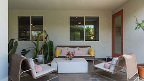 Ein überdachter Patio-Bereich mit einer Couch, zwei Korbstühlen, einem kleinen Tisch und Blumentöpfen.