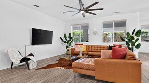 Ein modernes Wohnzimmer mit einer Ledersofa, einem Flachbildfernseher, einem Deckenventilator und großen Fenstern.