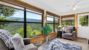 Ein Zimmer mit großen Fenstern und Blick auf eine bewaldete Landschaft, einem Deckenventilator und einer Sitzecke mit Sofa und Sessel.