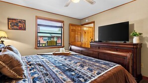 A bedroom with a large bed, a television, a wooden dresser, a window with blinds, and a door. - Dancing Bear by Avantstay Cozy Cabin + Hot Tub + Decks Arcade + Mountain Views Near Park (Sevierville)