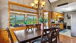 A dining area with a wooden table and chairs, a view of the outdoors through large windows, and a kitchen with wooden cabinets and a refrigerator. - Dancing Bear by Avantstay Cozy Cabin + Hot Tub + Decks Arcade + Mountain Views Near Park (Sevierville)