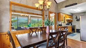A dining area with a wooden table and chairs, a view of the outdoors through large windows, and a kitchen with wooden cabinets and a refrigerator. - Dancing Bear by Avantstay Cozy Cabin + Hot Tub + Decks Arcade + Mountain Views Near Park (Sevierville)