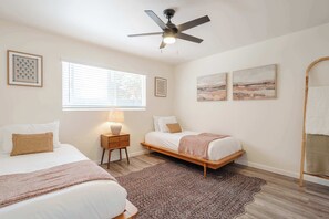 A bedroom with two beds, a ceiling fan, a window with blinds, and artwork on the walls. - Coastal 2BR | Walk to Ocean Beach & Cafés (San Diego)